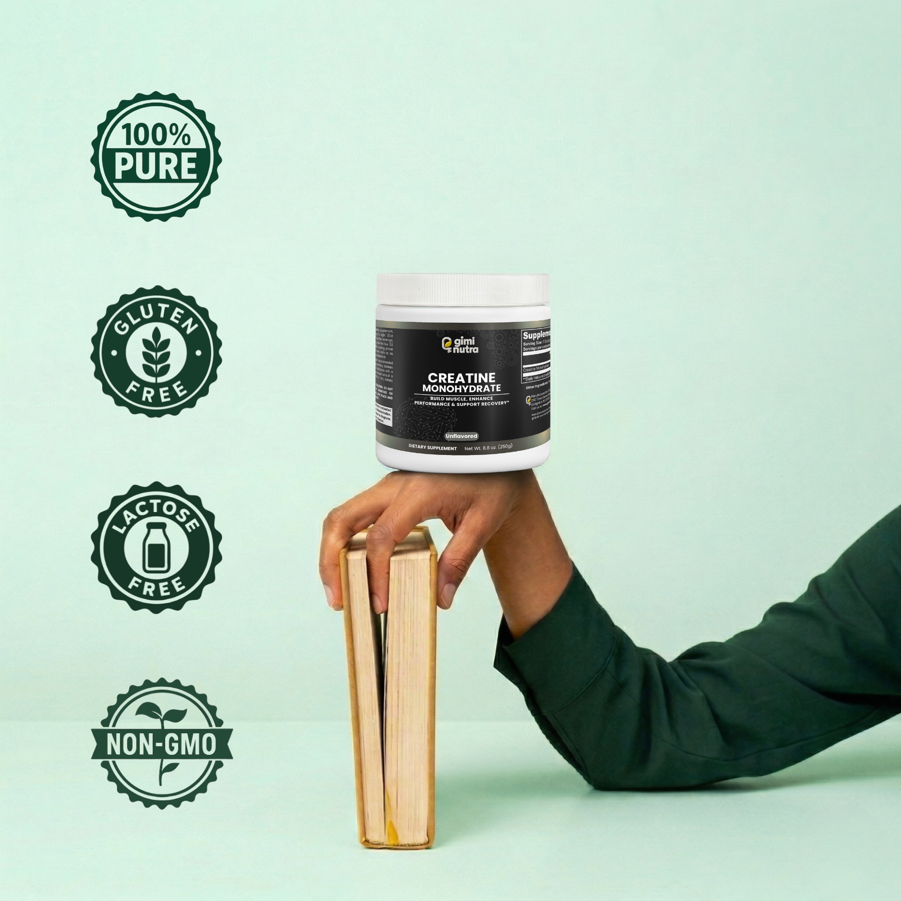 A person's arm resting on a mint green surface. The person is wearing a dark green long-sleeved and holding a thick, brownish book upright with their fingers. On top of their hand, giminutra creatine powder is balanced perfectly. Four trust badges are also to the left of the image. 