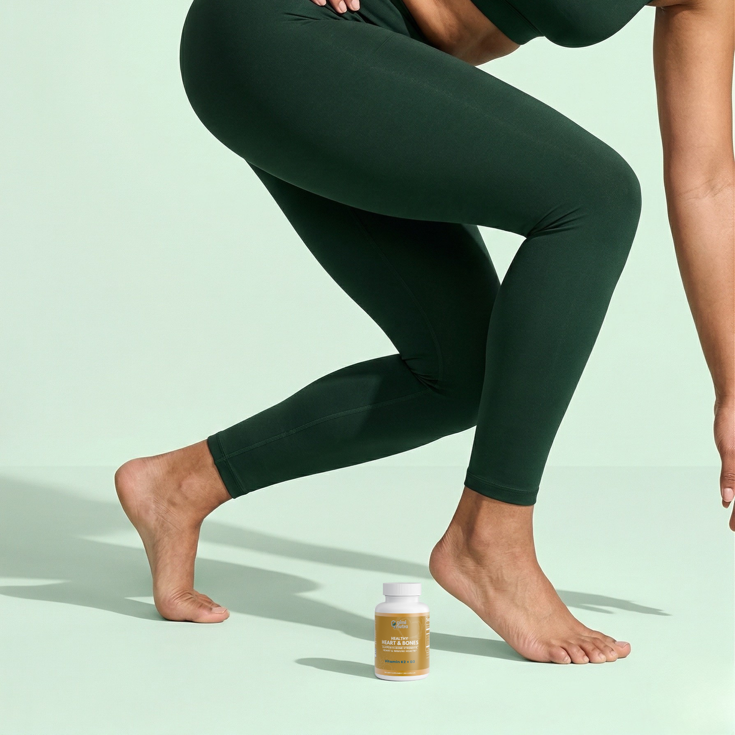 Person leaning down, wearing dark green leggings with a bottle of giminutra's vitamin K2+D3 supplement on a mint green background.