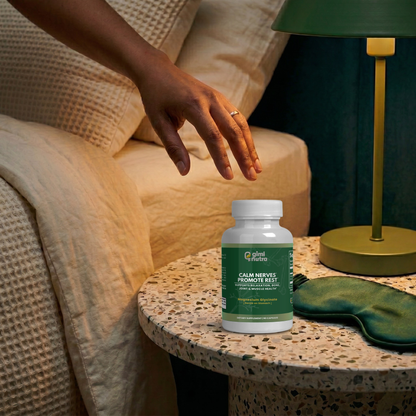 A warm, dimly lit bedroom scene featuring a Giminutra's magnesium glycinate supplement bottle on a terrazzo side table. A person's hand reaches down toward the bottle. Next to the bottle is a green silk sleep mask and an olive green bedside lamp, creating a relaxing nighttime atmosphere.