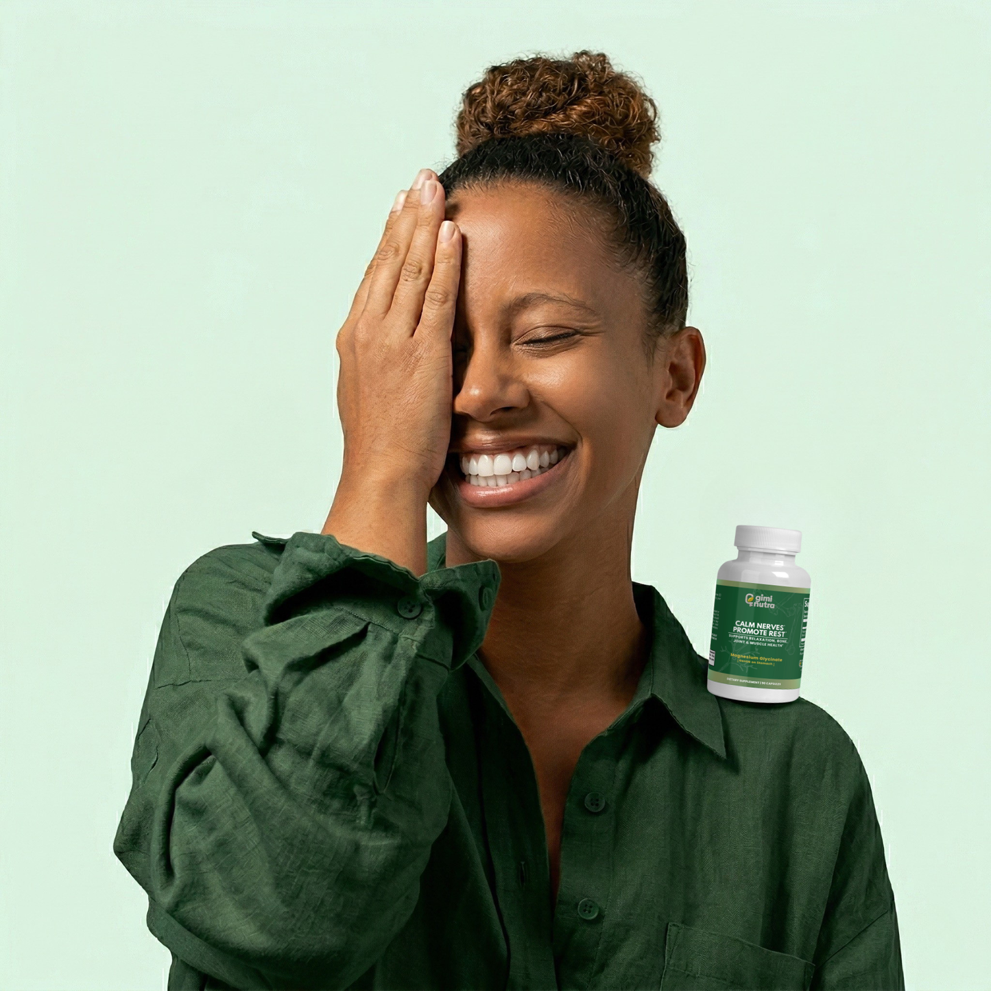 Woman happily smiling in green shirt with a bottle of giminutra magnesium glycinate on her shoulder on a mint green background