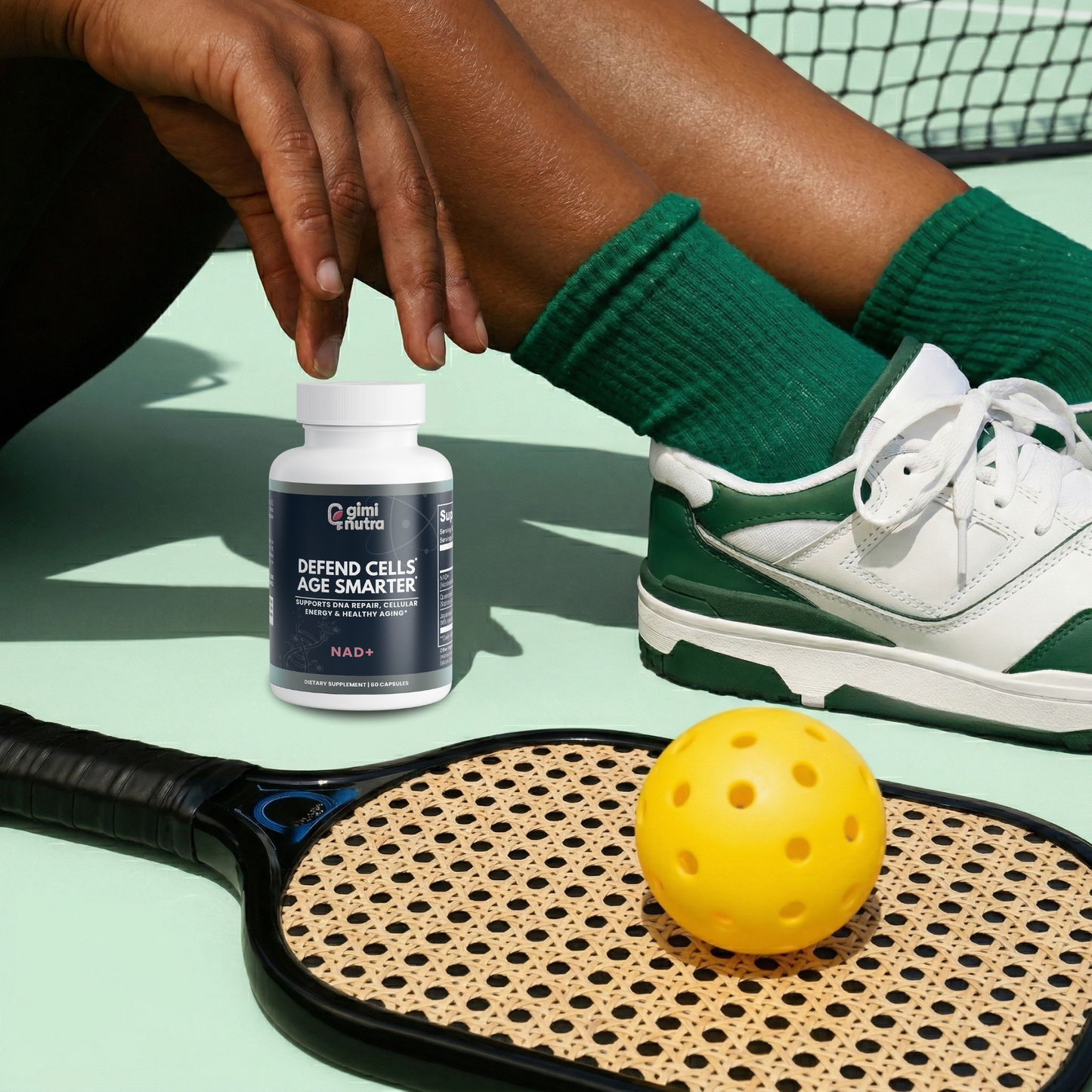 Person sitting on a pickleball court with a giminutra NAD+ supplement bottle and pickleball racket.