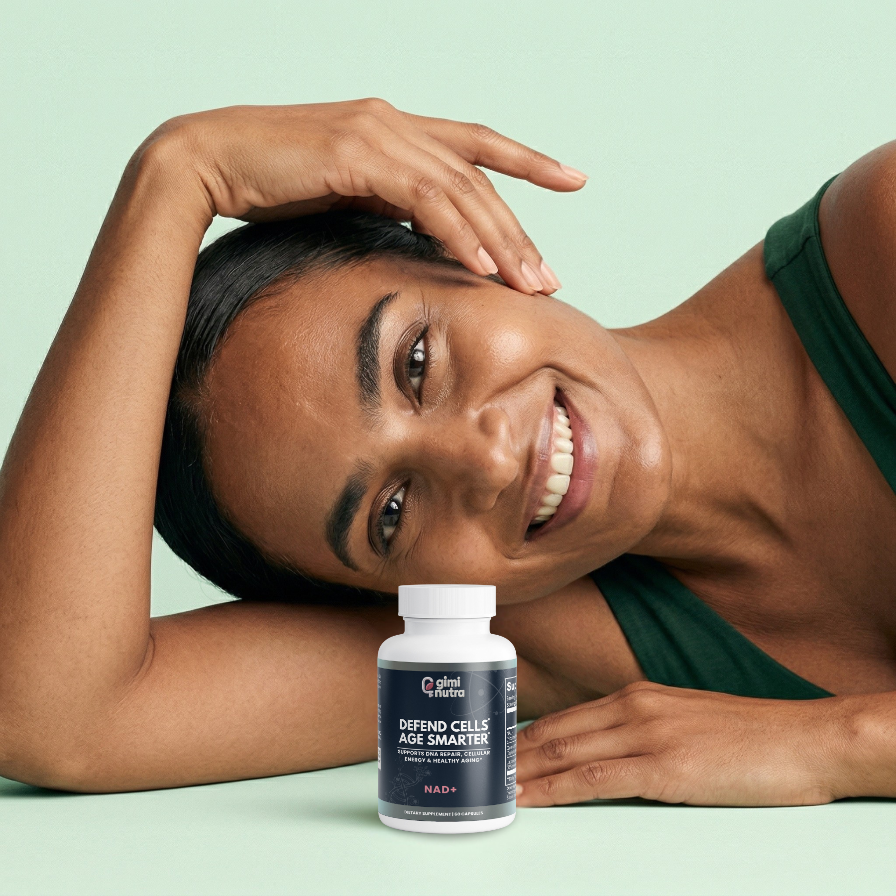 Youthful looking woman happily lying on a mint green background with a bottle of giminutra's NAD+ supplement bottle in front of her.